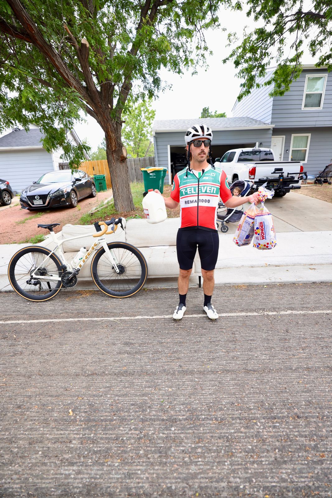 Carter Luense in a retro 7-Eleven jersey holding a gallon of milk and loaves of bread beside the white Trek at the curb