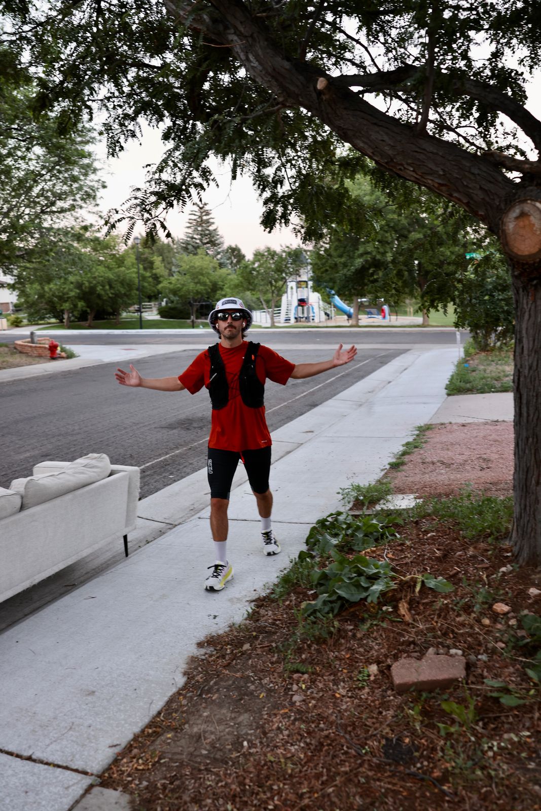 Carter Luense arms wide on the sidewalk under the tree, run vest and bucket hat